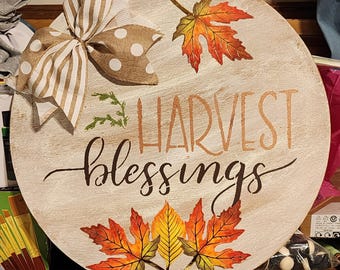May include: A round wooden sign with the words "Harvest Blessings" in brown lettering. The sign is decorated with a brown and white striped bow with white polka dots, and three fall-colored leaves.
