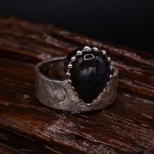 Puede incluir: Un anillo de plata con una piedra oscura en forma de lágrima. El anillo tiene una banda texturizada y un engaste decorativo alrededor de la piedra. La piedra es de un color oscuro, posiblemente negro o azul oscuro.