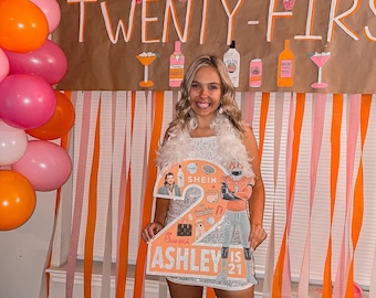 May include: A woman in a sparkly white dress holds a large pink and white number "2" decorated with images and text. The text on the sign reads "Ashley is 21". The background is decorated with pink and orange streamers and balloons.