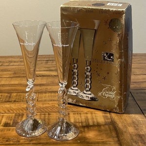 May include: Two clear glass champagne flutes with a twisted stem design. The flutes are in a box with the words "South Sea Resort" and "Cristal d'Arques" printed on it. The box also has the year 2000 printed on it.