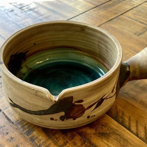May include: A brown ceramic bowl with a teal glaze inside and a brown handle. The bowl has a unique design with brown and black paint splatters.