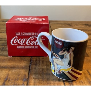 May include: A white ceramic mug with a Coca-Cola logo and a vintage illustration of a woman in a white swimsuit holding a bottle of Coca-Cola. The mug is 16 ounces and has a red box with the Coca-Cola logo and the text "Tasse en ceramique de 48 cl. Jarro de ceramica de 16oz."