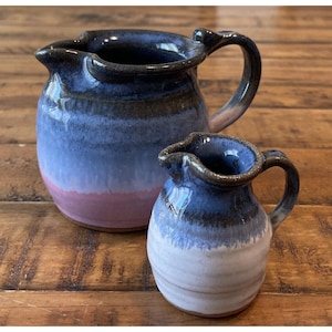 May include: Two ceramic pitchers, one larger than the other, with a blue, pink, and black glaze. The smaller pitcher is in front of the larger pitcher.