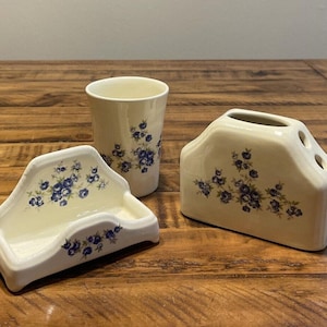 May include: A white ceramic bathroom set with a blue floral pattern. The set includes a toothbrush holder, a soap dish, and a tumbler.