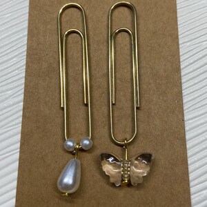 May include: Two gold paper clips with decorative charms. One clip has a white pearl teardrop and two smaller pearls. The other clip has a clear butterfly charm.