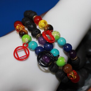 Puede incluir: Tres pulseras de cuentas con piedras preciosas de colores y cuentas de roca volcánica. Las pulseras presentan una variedad de colores, incluyendo rojo, amarillo, verde, azul y morado. Cada pulsera tiene un dije, incluyendo una forma geométrica roja y un diseño circular negro.