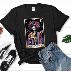 Puede incluir: Una camiseta negra de manga corta con un gráfico de un esqueleto que lleva una corona de flores y una túnica colorida, sosteniendo una taza de café. El diseño está enmarcado como una carta de tarot con el texto THE SPONSOR en la parte inferior.