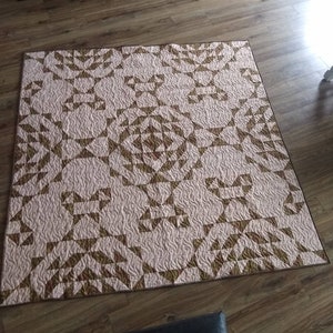 May include: A handmade quilt with a geometric pattern in shades of brown and pink. The quilt features a repeating design of triangles and other shapes, with a quilted texture. The quilt is rectangular and appears to be made of cotton.