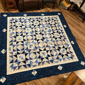 May include: A large, handmade quilt with a complex geometric pattern. The quilt features a central design of blue and cream-colored triangles and squares, framed by a dark blue border with diamond accents. The quilt is likely made of cotton.