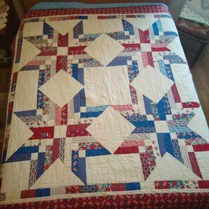 May include: A handmade quilt featuring a star pattern in red, white, and blue fabrics. The quilt has a white background with large star designs and a red border with a repeating pattern. The quilt is on a bed.