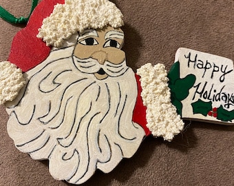 May include: A wooden Santa Claus ornament with a white beard and red hat. He is holding a sign that says "Happy Holidays" with green holly.