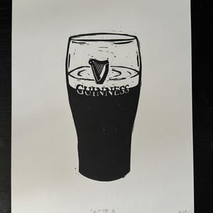 Puede incluir: Impresión en blanco y negro de una pinta de Guinness. El vaso está lleno de cerveza negra oscura, con el logotipo de Guinness y el símbolo del arpa. El texto "Split the G" está en la parte inferior.