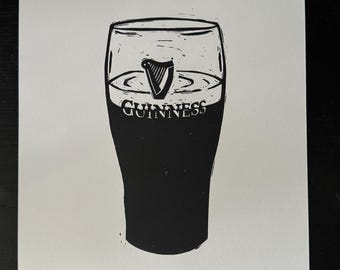 Puede incluir: Impresión en blanco y negro de una pinta de Guinness. El vaso está lleno de cerveza negra oscura, con el logotipo de Guinness y el símbolo del arpa. El texto "Split the G" está en la parte inferior.