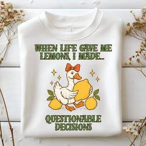 Op de afbeelding: Witte sweatshirt met een cartoon gans met een rode strik die een citroen vasthoudt. De tekst luidt "WHEN LIFE GAVE ME LEMONS, I MADE... QUESTIONABLE DECISIONS" in groene letters. Het ontwerp bevat gele citroenen en sterren.