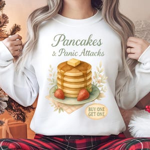 Può includere: Felpa bianca con la scritta "Pancakes & Panic Attacks" in verde. La grafica presenta una pila di pancake con burro e sciroppo, due fragole e elementi floreali. Un piccolo adesivo ovale dice "COMPRA UNO, PRENDI UNO".