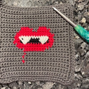 May include: A gray crocheted square with a red heart with white teeth and a black tongue in the center. A green crochet hook is lying on a gray surface next to the square.