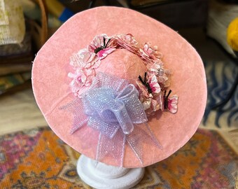 May include: A pink sun hat with a large brim, decorated with a floral trim and a light blue bow. The hat is on a white stand.