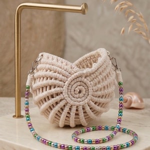 Puede incluir: Un bolso de macramé color crema con un patrón de nudos en espiral y una correa desmontable hecha de cuentas metálicas multicolores en tonos morado, azul, verde y dorado, sujeta con cierres de mosquetón de metal plateado.