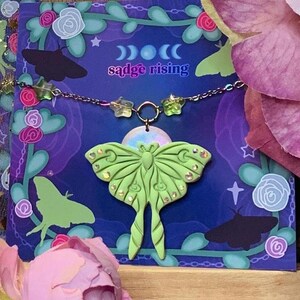 Peut inclure: Deux colliers avec des pendentifs en forme de papillon de nuit vert sur un fond violet avec des étoiles et des lunes dorées. Les colliers sont emballés dans un sac en organza transparent avec des étoiles et des lunes dorées. La carte indique "sadge rising".