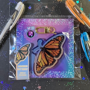 May include: A clear plastic bag containing a gold keychain with a monarch butterfly charm. The charm is a clear acrylic with a colorful butterfly design. The bag also contains a small card with the text "MONARCH BUTTERFLY" and "SAGITTARIUS".