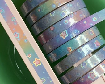 May include: A roll of iridescent washi tape with a rainbow gradient and a repeating pattern of pink stars. The tape is wound around a plastic spool.
