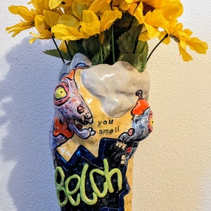 May include: A ceramic vase shaped like a cartoon monster with a yellow, orange, blue, and green paint scheme. The vase has three legs and the words "you smell" and "Belch" are painted on the front. The vase is filled with a bouquet of yellow sunflowers.