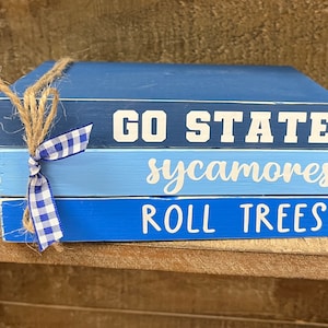 May include: A stack of three blue painted wooden blocks with white lettering. The top block reads "GO STATE", the middle block reads "sycamores", and the bottom block reads "ROLL TREES". The blocks are tied together with twine and a blue and white gingham bow.