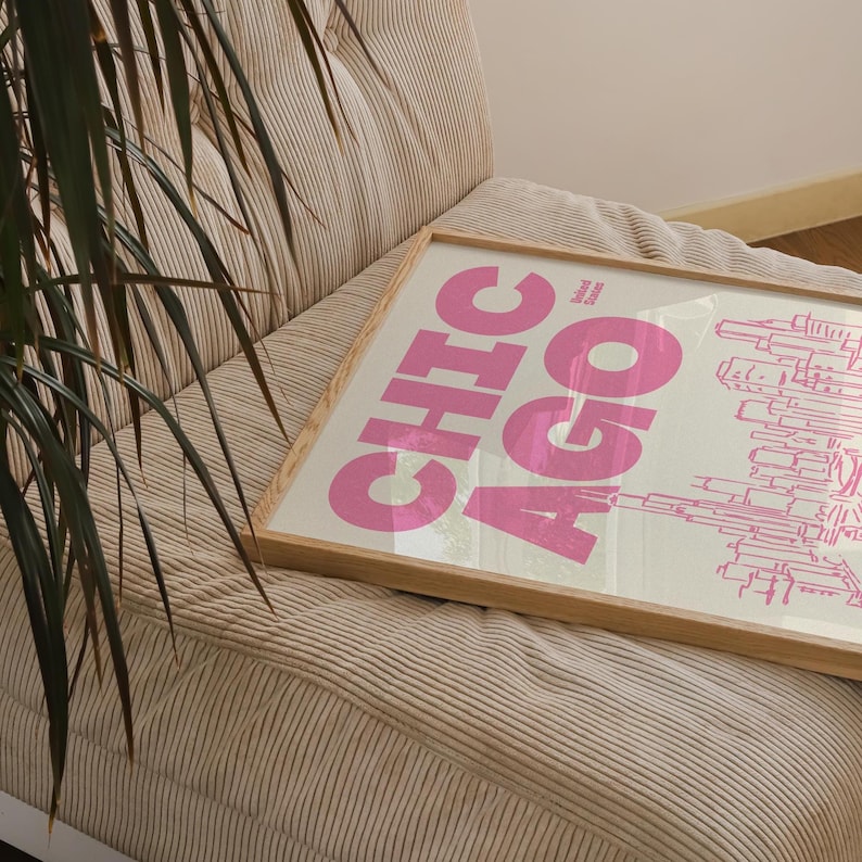 Op de afbeelding: Een ingelijste print met het woord CHICAGO in grote, dikke roze letters met een roze lijntekening van een stadsskyline. De tekst United States staat in kleine zwarte letters boven de skyline op een witte achtergrond.