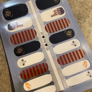May include: Nail art strips featuring designs inspired by a popular fantasy series. The strips include black, white, and burgundy nail decals with gold accents. Designs include a character with glasses and a scarf, a symbol, and striped patterns.
