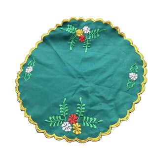 May include: A round, emerald green placemat with a scalloped gold border. The placemat features embroidered floral designs in red, yellow, and white. The embroidered leaves are green.