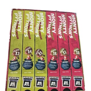May include: A collection of six Monty Python's Flying Circus VHS tapes, volumes 13 through 18, arranged in a box set. The first three volumes have light green spines, and the last three have red spines, each featuring a cartoon illustration.
