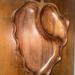 Puede incluir: Una bandeja de madera con forma de hoja con dos compartimentos y un asa. La bandeja está hecha de madera marrón oscuro con un patrón de vetas natural. Los bordes de la bandeja tienen un diseño ondulado.