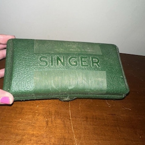 May include: A rectangular, dark green, textured plastic storage case with the word SINGER embossed in capital letters on the front. The case features a hinged design with a central latch mechanism and a pebbled surface texture throughout the exterior.