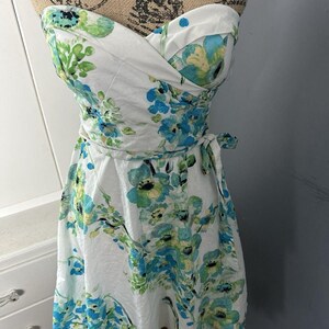 Puede incluir: Un vestido blanco sin tirantes con un estampado floral en tonos azules y verdes. El vestido presenta un corpiño ajustado y una falda acampanada, con una cinta en la cintura. El vestido se exhibe en un maniquí.