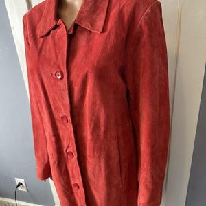 May include: A red suede jacket with a collared neckline and button-down closure. The jacket has long sleeves and a tailored fit. The color is a vibrant, warm red, and the suede texture is visible. The jacket is displayed on a mannequin.