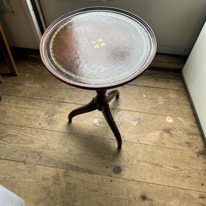 May include: A round wooden pedestal table with a dark brown finish. The tabletop features a circular leather inlay with a decorative gold-colored border and a central gold-colored emblem. The table stands on a single central column with three legs.