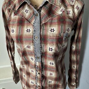 Puede incluir: Una camisa abotonada de manga larga con un estampado de cuadros rojos, marrones y beige. La camisa presenta costuras decorativas y un acento a rayas en la parte delantera. El cuello y los puños también son a cuadros. La camisa tiene un estilo occidental.