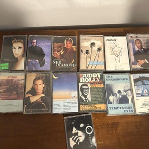 May include: A collection of vintage cassette tapes featuring various artists. The tapes showcase album covers with portraits, graphics, and text. The collection includes artists like Neil Diamond, Celine Dion, and Buddy Holly.