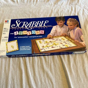Pode incluir: Uma caixa do jogo de tabuleiro Scrabble Juniors com fundo azul e o título do jogo em letras brancas e vermelhas. A caixa apresenta uma imagem de duas crianças jogando. A caixa também inclui o texto "Tabuleiro de 2 lados para iniciantes e avançados!"