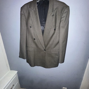 Könnte beinhalten: Ein zweireihiger Blazer mit braun-schwarzem Hahnentrittmuster. Das Kleidungsstück verfügt über breite Revers, zwei Pattentaschen an der Taille und dunkle Knöpfe. Das Innere ist mit schwarzem Stoff gefüttert und hängt an einem Kunststoffbügel.