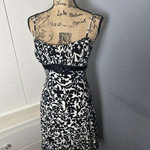 Puede incluir: Vestido estampado en blanco y negro con tirantes finos y un lazo negro en la cintura. El vestido se exhibe en un maniquí. El estampado presenta un diseño floral. El vestido tiene una falda acampanada.