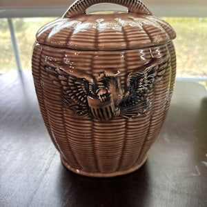 Könnte beinhalten: Braune Keramik-Keksdose mit Deckel und Griff. Die Dose hat ein Korbgeflecht-Design und ein geprägtes Adleremblem. Der Deckel hat ebenfalls einen Griff. Die Dose steht auf einer dunkelbraunen Oberfläche.
