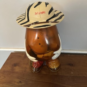 Puede incluir: Figura de cerámica de una persona con sombrero de safari con la palabra "Zookeeper" impresa. El sombrero tiene un estampado de rayas de cebra. La figura es marrón con guantes blancos, zapatos marrones y una pajarita roja.