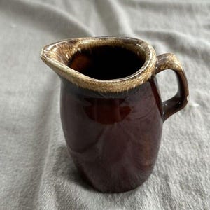 Può includere: Una piccola lattiera in ceramica con una smaltatura lucida color marrone scuro. Il bordo presenta uno smalto a contrasto in stile gocciolato in una tonalità beige più chiara. La brocca ha un corpo arrotondato, un piccolo beccuccio e un manico.