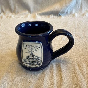 Puede incluir: Una taza de cerámica azul oscuro con cuerpo redondeado y asa curva. Una etiqueta rectangular de color crema en la parte frontal presenta un dibujo lineal negro de una casa histórica y el texto 1884 PHIPPS INN Hudson.