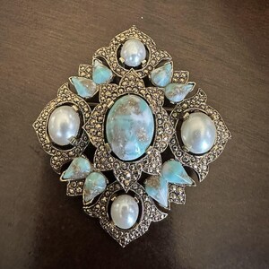 May include: Ornate, vintage-style brooch with a central oval turquoise stone, surrounded by faux pearls and smaller turquoise accents. The brooch has a detailed, gold-toned metalwork design, creating a luxurious and eye-catching accessory.