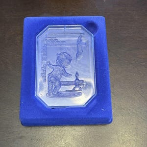 May include: A blue glass plaque with a child-like engraving, set in a blue velvet box. The engraving depicts a child playing near a water fountain. The plaque has an octagonal shape.