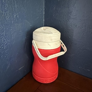 May include: A red plastic beverage cooler with a white screw-top lid and a white swinging handle. The cylindrical container features a molded, contoured body and a textured base. The word Igloo is embossed in raised letters around the bottom edge.