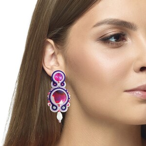 May include: Purple and pink statement earrings with a white pearl accent. The earrings are made with colorful thread and beads.