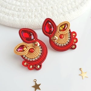 以下が含まれることがあります： 幾何学模様の赤と金のイヤリングのペア。イヤリングは糸でできており、赤と金のビーズがいくつか付いています。